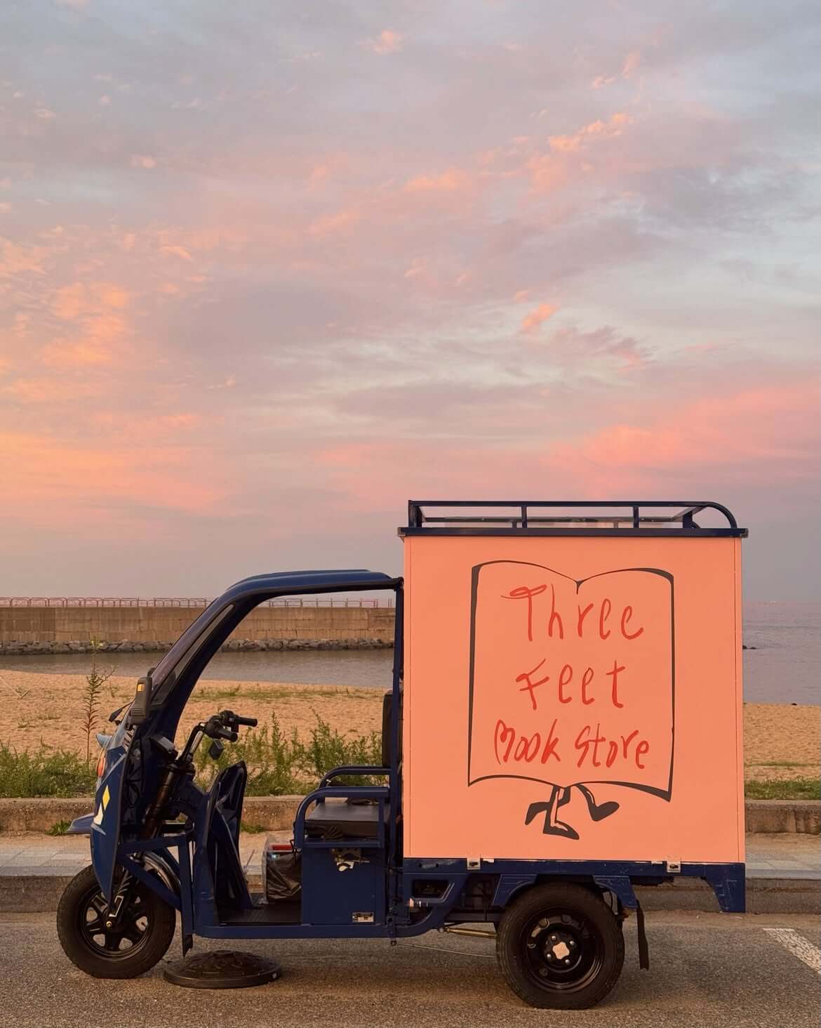 寫著 Three Feet Bookstore 的三輪書車停在海岸邊,天空帶有粉橘色晚霞。